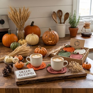May include: A wooden serving board holds two white mugs with latte art, resting on orange and mauve crochet pumpkin coasters. A small sign reads HELLO AUTUMN. Nearby, a gift box contains four additional crochet pumpkin coasters in various colors.