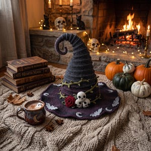 Pode incluir: Um chapéu de bruxa de crochê roxo escuro com uma ponta alta e curva, aba larga e elementos decorativos, incluindo uma trepadeira verde, uma rosa vermelha, três pequenas caveiras brancas e motivos de morcegos e luas na borda da aba.