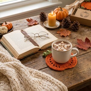 May include: A white ceramic mug filled with hot cocoa and star-shaped marshmallows sits on an orange crocheted pumpkin coaster. Nearby is an open book with round glasses, a lit yellow pillar candle, pinecones, small pumpkins, and autumn leaves.