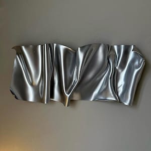 Könnte beinhalten: Eine rechteckige Wandskulptur aus silberfarbenem Metall mit einer Reihe tiefer, fließender Falten und Kurven, die ein welliges, stoffartiges Aussehen erzeugen, mit glatten, reflektierenden Oberflächen und deutlichen Schatten auf der Metallform.