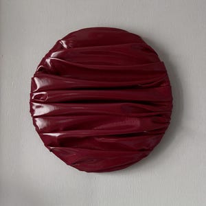 May include: A circular wall art piece featuring a deep burgundy, high-gloss finish. The surface is characterised by horizontal, undulating folds and ridges that create a layered, fabric-like appearance with reflective highlights across the curves.
