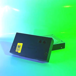 May include: A black rectangular network adapter featuring two phone ports and a yellow warning label that reads DO NOT CONNECT PHONE LINE. Behind it sits a silver hard drive. The device has a smooth plastic casing with visible connection ports.