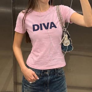 May include: A light pink short-sleeved t-shirt with the word DIVA printed in dark blue letters across the chest, paired with loose-fitting blue denim jeans featuring a distressed knee hole and a small dark blue shoulder bag with silver accents.