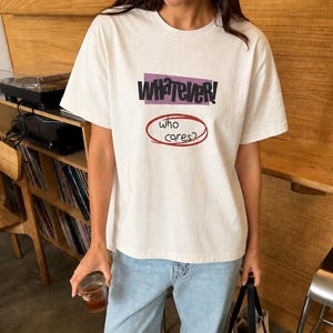 Może przedstawiać: Biały t-shirt z krótkim rękawem z napisem "whatever!" w kolorze czarnym na fioletowym prostokącie oraz napisem "who cares?" w kolorze czarnym wewnątrz czerwonego owalu. Koszulka zestawiona jest z jasnoniebieskimi dżinsami, czarnymi mokasynami i beżową torbą.