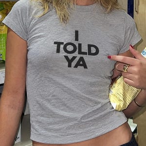 May include: A heather grey short-sleeved t-shirt featuring the black text I TOLD YA printed on the chest. The garment has a crew neckline and a fitted silhouette, constructed from a soft, lightweight fabric with a visible heathered texture.