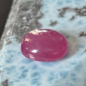 Puede incluir: Una piedra preciosa de color rosa vibrante, lisa y de forma ovalada, que descansa sobre una losa triangular pulida de piedra azul claro y blanca con inclusiones de color gris oscuro, moteadas y texturizadas en toda su superficie.