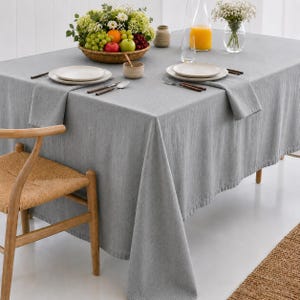 Könnte beinhalten: Eine rechteckige graue Leinentischdecke auf einem Esstisch, gedeckt mit zwei weißen Keramiktellern, Besteck, einer kleinen Schale, einer Glaskaraffe mit Orangensaft, einem geflochtenen Obstkorb und einer Vase mit weißen Blumen.