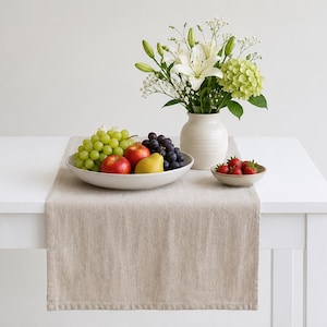 May include: A beige linen table runner draped over a white table. A large white bowl holds green grapes, red apples, a yellow pear, and dark grapes. A white vase contains white lilies and green hydrangeas, and a small bowl holds red strawberries.