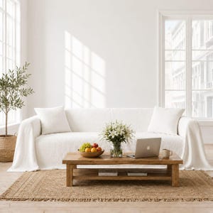 May include: A white sofa with two matching throw pillows, a wooden coffee table holding a bowl of fruit, a vase of white lilies, and a silver laptop. A woven rug sits underneath the table, and a potted olive tree stands to the left.