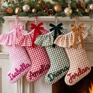 Puede incluir: Cuatro calcetines navideños de tela con estampado de cuadros vichy en rosa, rojo, verde y beige. Cada calcetín tiene un borde con volantes, un lazo de terciopelo y un nombre bordado en cursiva: Isabella, Amelia, Diana y Jordan.