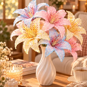 May include: A bouquet of artificial lily flowers in pastel shades of blue, pink, and yellow with speckled patterns, arranged in a white ceramic vase with a spiraled, twisted design. The lilies feature prominent stamens and open, flared petals.