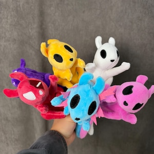 May include: A collection of six small plush toys in various colours including yellow, white, blue, pink, red, and purple. Each plush features large black oval eyes, pointed ears, and a soft, fuzzy texture. The toys are grouped together in a cluster.