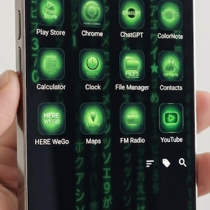 Può includere: Uno smartphone con cornice metallica mostra un'interfaccia digitale con sfondo scuro e caratteri giapponesi verdi verticali. Lo schermo presenta una griglia di icone di app verde brillante, tra cui Play Store, Chrome, ChatGPT e altre ancora.