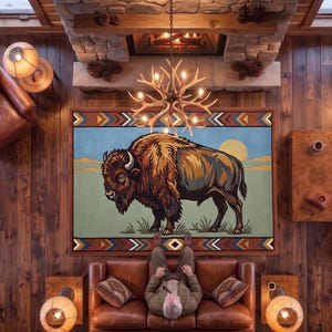 May include: A rectangular area rug featuring a large, detailed illustration of a brown bison standing in a grassy field under a yellow sun, framed by a geometric border with blue, orange, and white triangular patterns on a dark background.