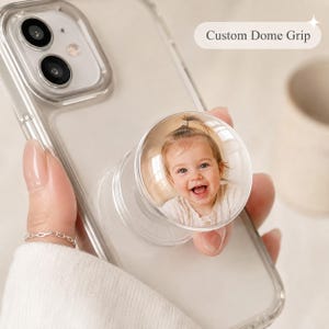 May include: A clear smartphone case with a circular custom dome grip attached to the back. The grip features a photographic print of a smiling baby. The text Custom Dome Grip is visible in the upper right corner of the image.