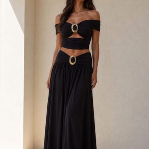 May include: A black two-piece set consisting of an off-the-shoulder crop top and a long, flowing skirt. Both pieces feature a central gold-toned oval hardware accent. The top has a cutout detail, and the skirt has a gathered waistband.