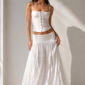 May include: A white two-piece set consisting of a sleeveless corset-style top with thin straps and a front button closure, paired with a long, tiered maxi skirt featuring a wide, smocked elastic waistband and a frayed hemline detail.