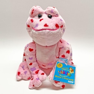 Puede incluir: Un juguete de peluche de rana en color rosa claro con un patrón de corazones rojos y morados. La rana tiene grandes ojos negros y una barriga rosa pálido. En su brazo lleva una etiqueta azul con el texto Webkinz discover a virtual world GANZ.