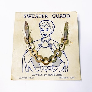 Puede incluir: Protector de suéter vintage en una tarjeta crema con una ilustración azul de una mujer. El protector tiene dos clips ornamentados conectados por una cadena de anillos color latón. El texto en la tarjeta dice "SWEATER GUARD" y "JEWELS by JEWELINE."