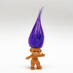 May include: A vintage troll doll with a tan body and a large, vibrant purple hair. The doll has a smiling expression and outstretched arms, a classic toy from the 1960s.