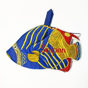 May include: Embroidered fish-shaped ornament in blue, yellow, and red. The design features a blue body with yellow stripes, a yellow tail, and a red head. The word "Saipan" is embroidered in red across the body. A blue loop is attached for hanging.