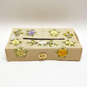 Pode incluir: Uma capa retangular para caixa de lenços de tecido bege tecido, adornada com flores bordadas em amarelo, branco e roxo. A capa tem um fecho dourado e uma abertura retangular para dispensar lenços.