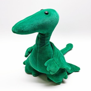 May include: A plush toy shaped like a green dinosaur with a long neck, a pointed snout, and black bead eyes. The creature features a soft, velvet-like texture, small front limbs, and a long tail extending to the right side of the frame.