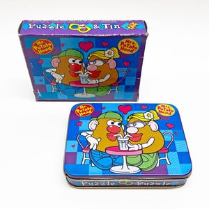 May include: A vintage Mr. and Mrs. Potato Head puzzle and tin set. The box and tin feature a cartoon illustration of the couple sharing a drink. The box is purple with the text "Puzzle & Tin". The tin is blue with the text "Puzzle".