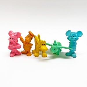 Könnte beinhalten: Eine Sammlung bunter Plastikfiguren mit Zeichentrickfiguren. Die Figuren umfassen eine rosa Maus, eine orangefarbene Ente, einen gelben Hund, ein grünes Flugzeug und eine blaue Maus. Die Spielzeuge stehen auf einer weißen Oberfläche.