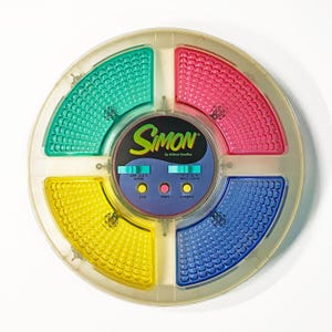 Może przedstawiać: Zabytkowa gra Simon firmy Milton Bradley. Okrągła gra ma cztery kolorowe kwadranty: zielony, czerwony, żółty i niebieski. Środek zawiera logo Simon i elementy sterujące grą. Gra jest przeznaczona do zapamiętywania i rozpoznawania wzorów.