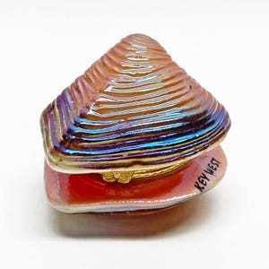 May include: Iridescent clam shell trinket box with a hinged lid. The shell features a textured surface with shades of orange, blue, and purple. The base is red with the text "KEY WEST". A gold-colored clasp secures the lid.
