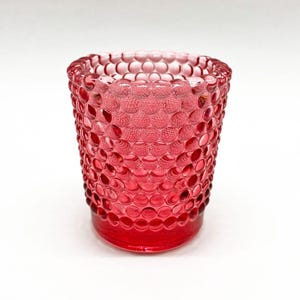 May include: A translucent red glass votive holder featuring a repeating hobnail pattern of raised circular bumps across the entire exterior surface. The vessel has a cylindrical shape with a slightly wider rim and a smooth, flat base.