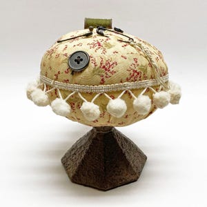 May include: A vintage-style pincushion with a floral pattern in red and green on a cream background. It features a large button, decorative trim, and white pom-poms. The pincushion sits on a dark brown, geometric base.