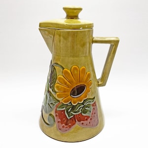 Puede incluir: Jarra de cerámica con tapa, de color amarillo verdoso apagado. La jarra presenta un diseño en relieve de girasol y fresas. El asa es angular y la jarra tiene un pico. La jarra es probablemente para servir bebidas.