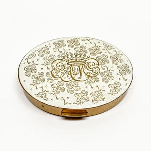 Pode incluir: Um estojo compacto oval vintage com uma parte superior em esmalte branco e uma base de metal dourado. A parte superior apresenta uma coroa dourada e um design de monograma, rodeado por um padrão repetido de laços e redemoinhos. O estojo compacto está fechado.