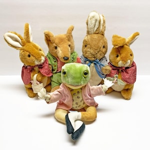 Könnte beinhalten: Eine Gruppe von Plüschtier-Spielzeugen, darunter Kaninchen, ein Fuchs und ein Frosch, sind vor einem weißen Hintergrund angeordnet. Die Kaninchen-Spielzeuge tragen rote Umhänge und kleine Körbe. Der Fuchs trägt eine grüne Jacke, und der Frosch eine rosa Jacke.