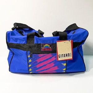 Puede incluir: Una bolsa de lona Gitano azul claro con asas y detalles en negro. La bolsa presenta un panel frontal con un diseño abstracto colorido en rosa, amarillo y morado. Se adjunta una etiqueta con el logotipo de Gitano.