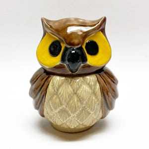 Op de afbeelding: Een keramieken uilfiguur met een bruin lichaam, gele ogen en een zwarte snavel. De uil heeft een gedetailleerd veermotief op zijn borst en vleugels. Het beeldje is een decoratief item.