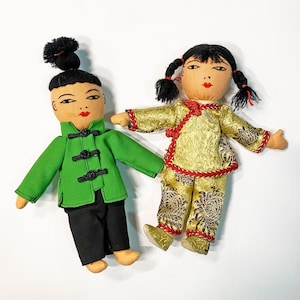 Puede incluir: Dos muñecas asiáticas vintage. Una muñeca viste una chaqueta verde y pantalones negros. La otra muñeca está vestida con un atuendo dorado con un estampado floral y ribetes rojos. Ambas muñecas tienen cabello negro y rasgos faciales pintados.