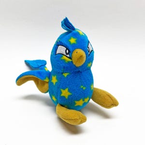 Puede incluir: Un juguete de peluche de pájaro azul con patrones de estrellas amarillas. El pájaro tiene pico, alas y patas amarillas. Tiene detalles blancos alrededor de los ojos y una cresta azul. El juguete está sobre una superficie blanca.