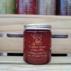 Può includere: Un barattolo di marmellata Crafted House Habanero Gold, peso netto 340g. La marmellata è di colore rosso intenso con scaglie di peperoncino visibili. Il barattolo ha un coperchio argentato ed è esposto su una cassa di legno.
