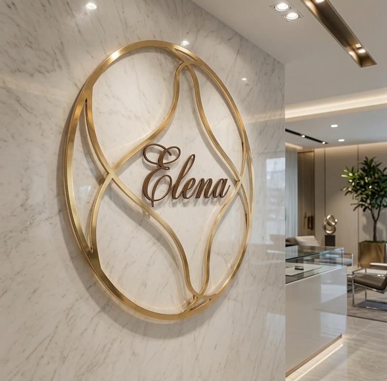 May include: A circular gold metal wall sign featuring an intricate, overlapping wire frame design. The word Elena is written in a dark, cursive script font in the center of the gold frame. The sign is mounted on a white marble wall with grey veining.