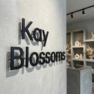 May include: A three-dimensional wall sign featuring the black text Kay Blossoms mounted on a light gray concrete surface. The letters are spaced slightly from the wall, creating distinct shadows that emphasize the depth of the signage.