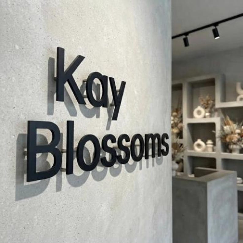 May include: A three-dimensional wall sign featuring the black text Kay Blossoms mounted on a light gray concrete surface. The letters are spaced slightly from the wall, creating distinct shadows that emphasize the depth of the signage.