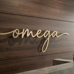 May include: A gold-colored metal sign featuring the word omega in a cursive script font. The lettering is mounted on a dark brown horizontal wood-paneled wall, creating a dimensional effect with a visible shadow cast behind the letters.