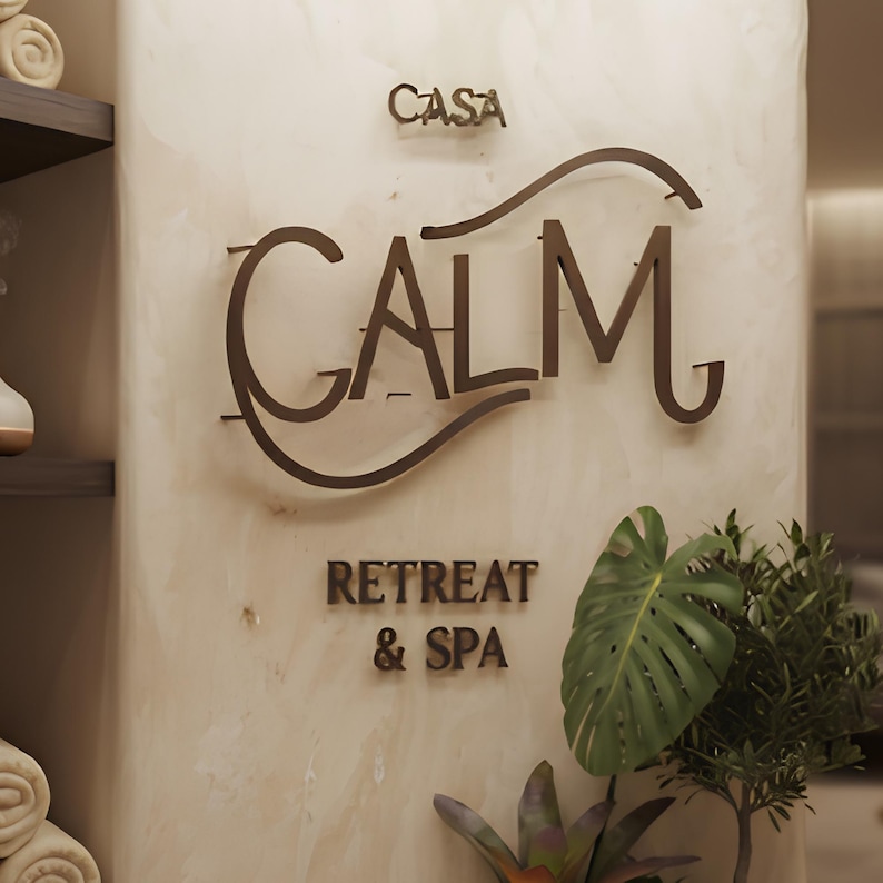 May include: A wall-mounted sign featuring the text CASA CALM RETREAT & SPA in dark brown, stylized lettering. The sign is mounted on a smooth, light beige, marbled surface. A green monstera leaf and other foliage are visible at the bottom right.