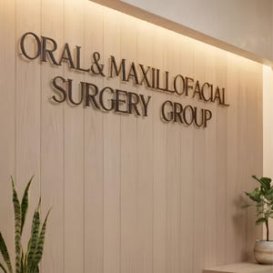 May include: A wall-mounted sign featuring three-dimensional dark metal lettering that reads ORAL & MAXILLOFACIAL SURGERY GROUP. The sign is mounted on a light-colored, vertical wood-paneled wall with integrated warm lighting at the top edge.