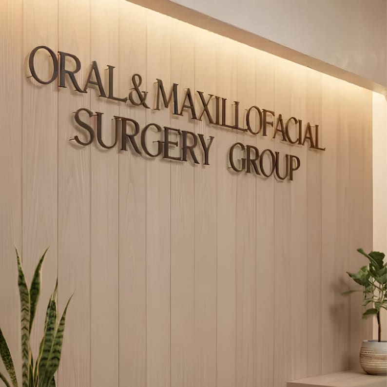 May include: A wall-mounted sign featuring three-dimensional dark metal lettering that reads ORAL & MAXILLOFACIAL SURGERY GROUP. The sign is mounted on a light-colored, vertical wood-paneled wall with integrated warm lighting at the top edge.