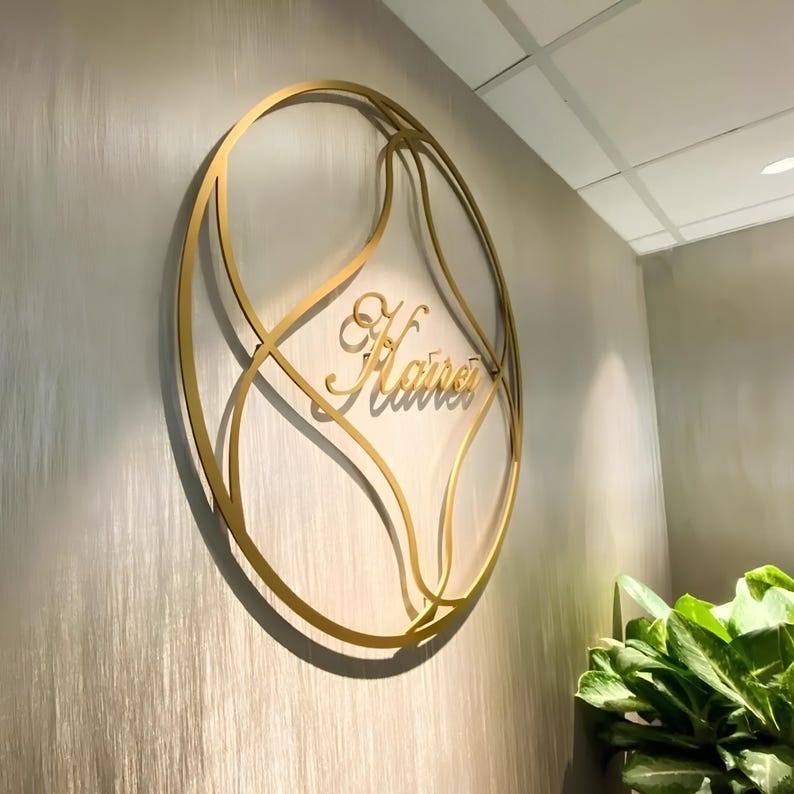 May include: A circular gold-colored metal wall sign featuring an intricate, flowing geometric frame. The center of the sign displays the word "Kairei" in a cursive, script-style font. The sign is mounted on a light-colored, vertically textured wall.