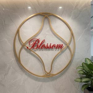 May include: A circular gold metal wall sign featuring an intricate, symmetrical geometric frame. The word Blossom is written in a red, cursive script font across the center of the frame. The sign is mounted on a light gray marble wall surface.